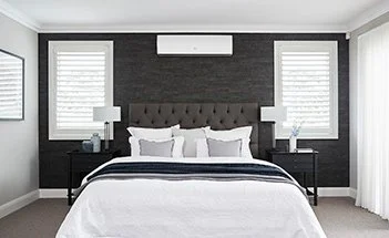A modern bedroom with a white bed, black headboard, two black nightstands with lamps, and windows with white blinds.