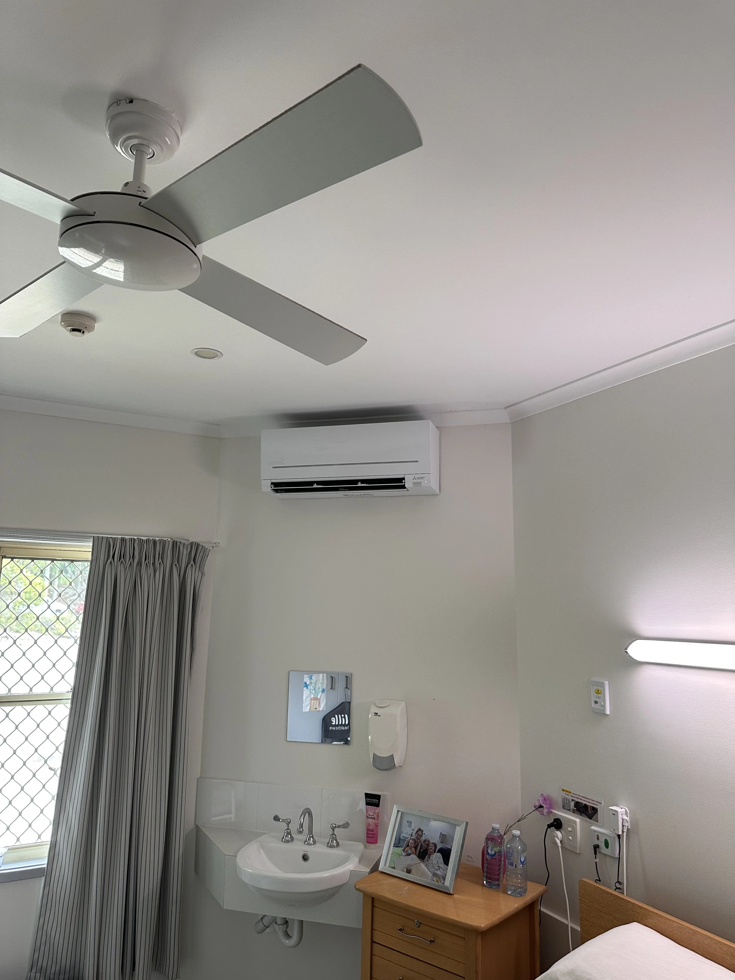 Hospital or healthcare room with ceiling fan, air conditioner, sink, mirror, and various personal items near a bed.