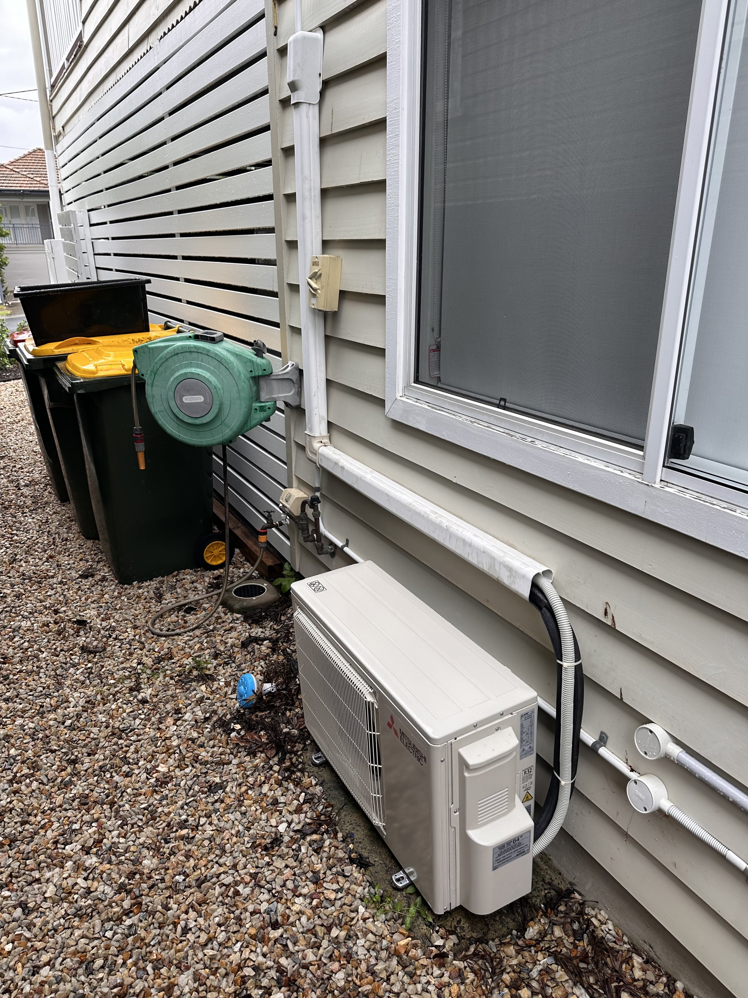 Exterior of a house with horizontal siding, a window with a screen, and various HVAC system units and wires attached to the wall, including a green retractable garden hose reel, a black trash bin, and a white Mitsubishi air conditioning unit on the g
