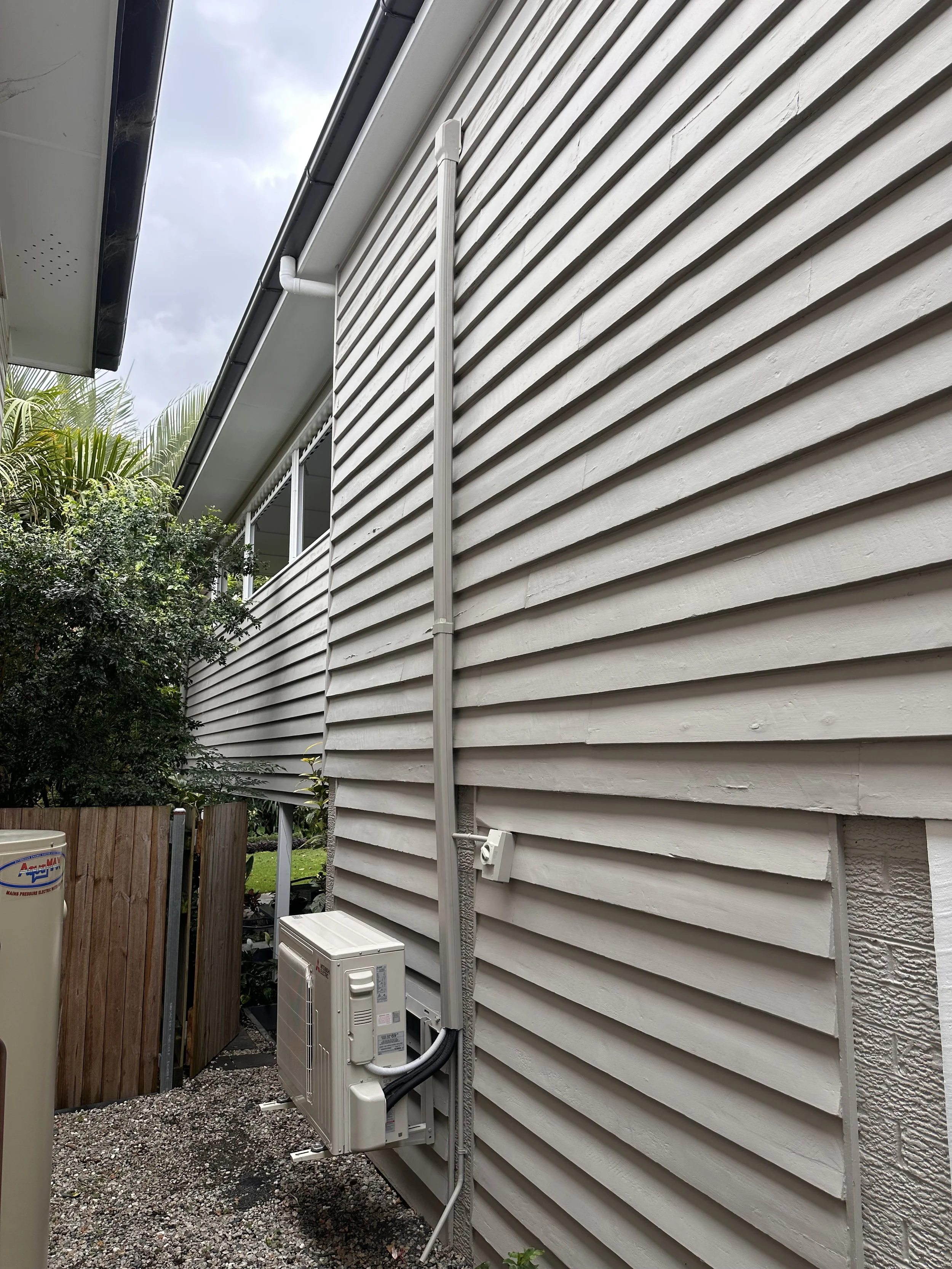 The side of a house with beige horizontal siding, showing a white metal pipe running vertically, an electrical box below, and an outdoor air conditioning unit. There are trees and a cloudy sky in the background.