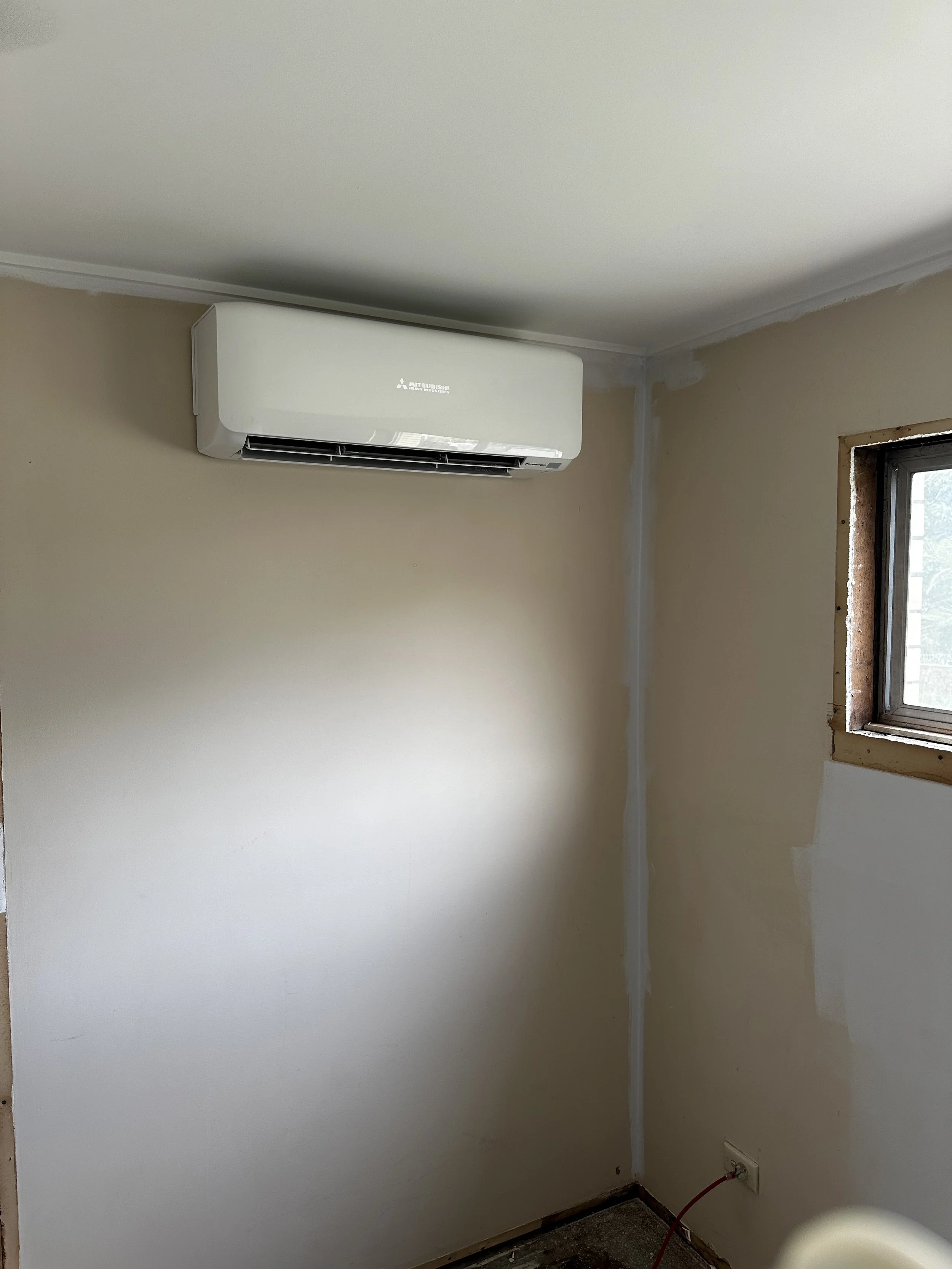 A room with beige walls, an air conditioning unit mounted near the ceiling, and an unfinished window frame with visible insulation and a power outlet with a red extension cord plugged in.
