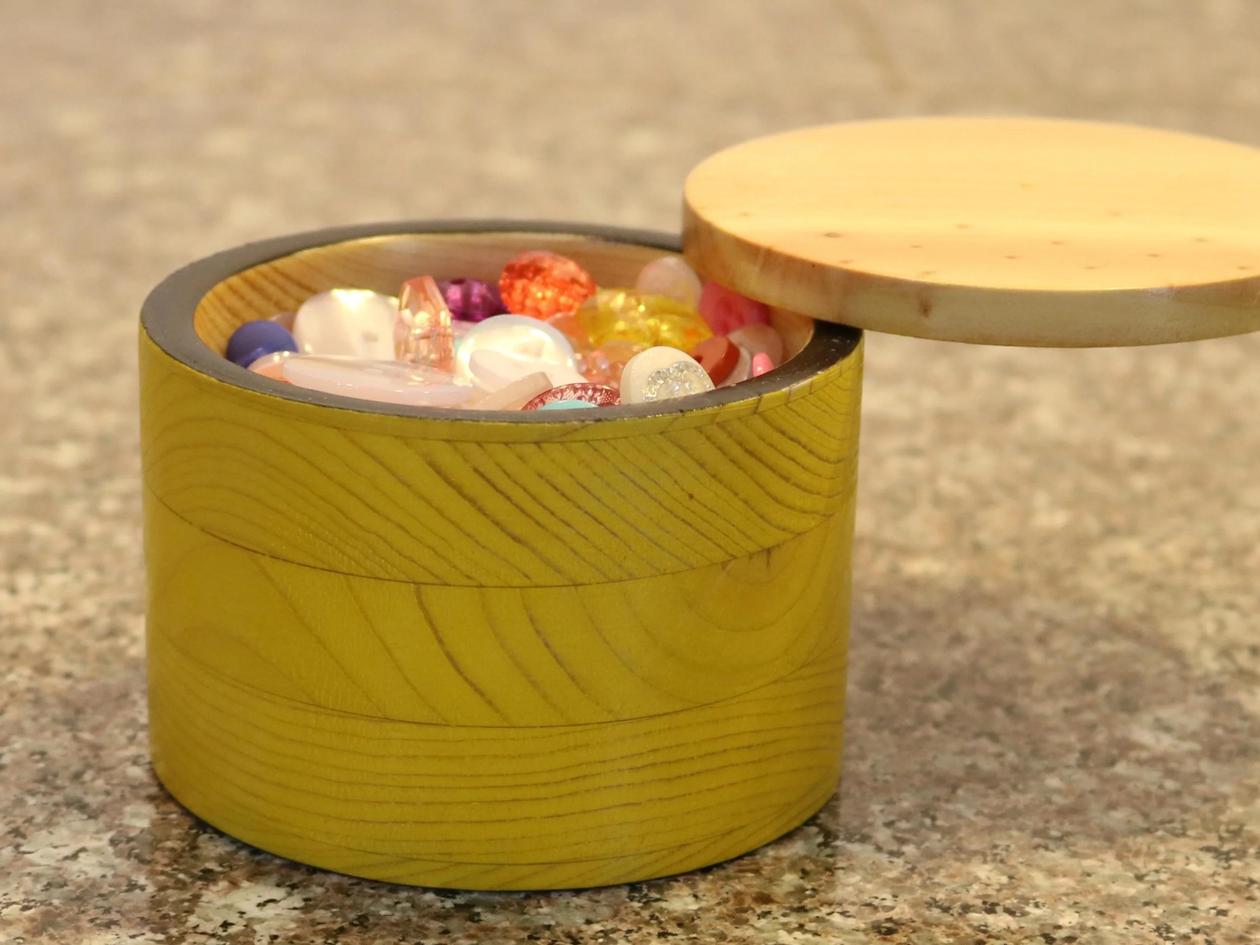 Yellow wooden container with its lid open, filled with assorted colorful glass marbles, placed on a speckled countertop.