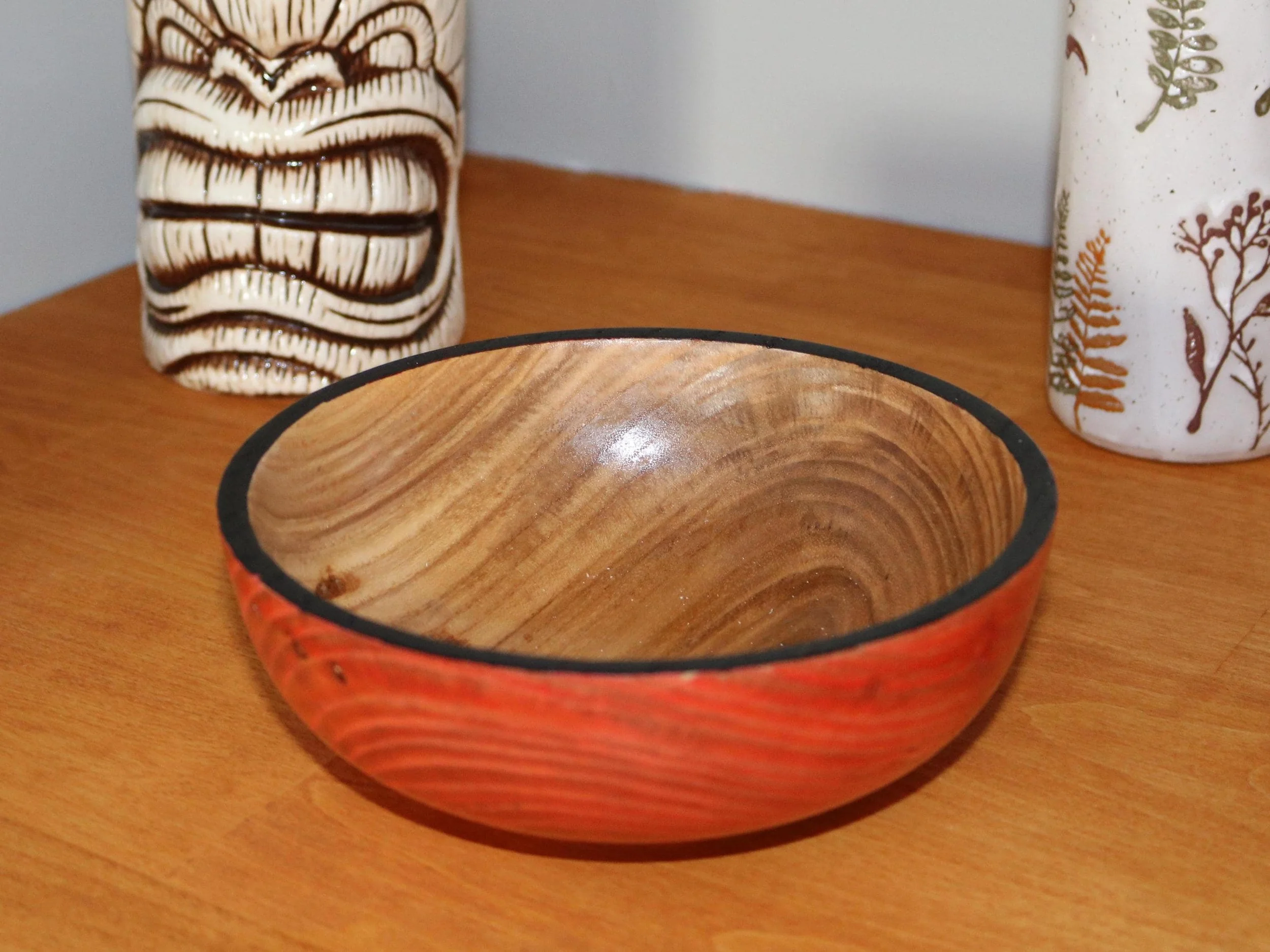A wooden bowl with a reddish exterior and a natural wood interior, flanked by two ceramic vases with nature-inspired patterns on a wooden surface.