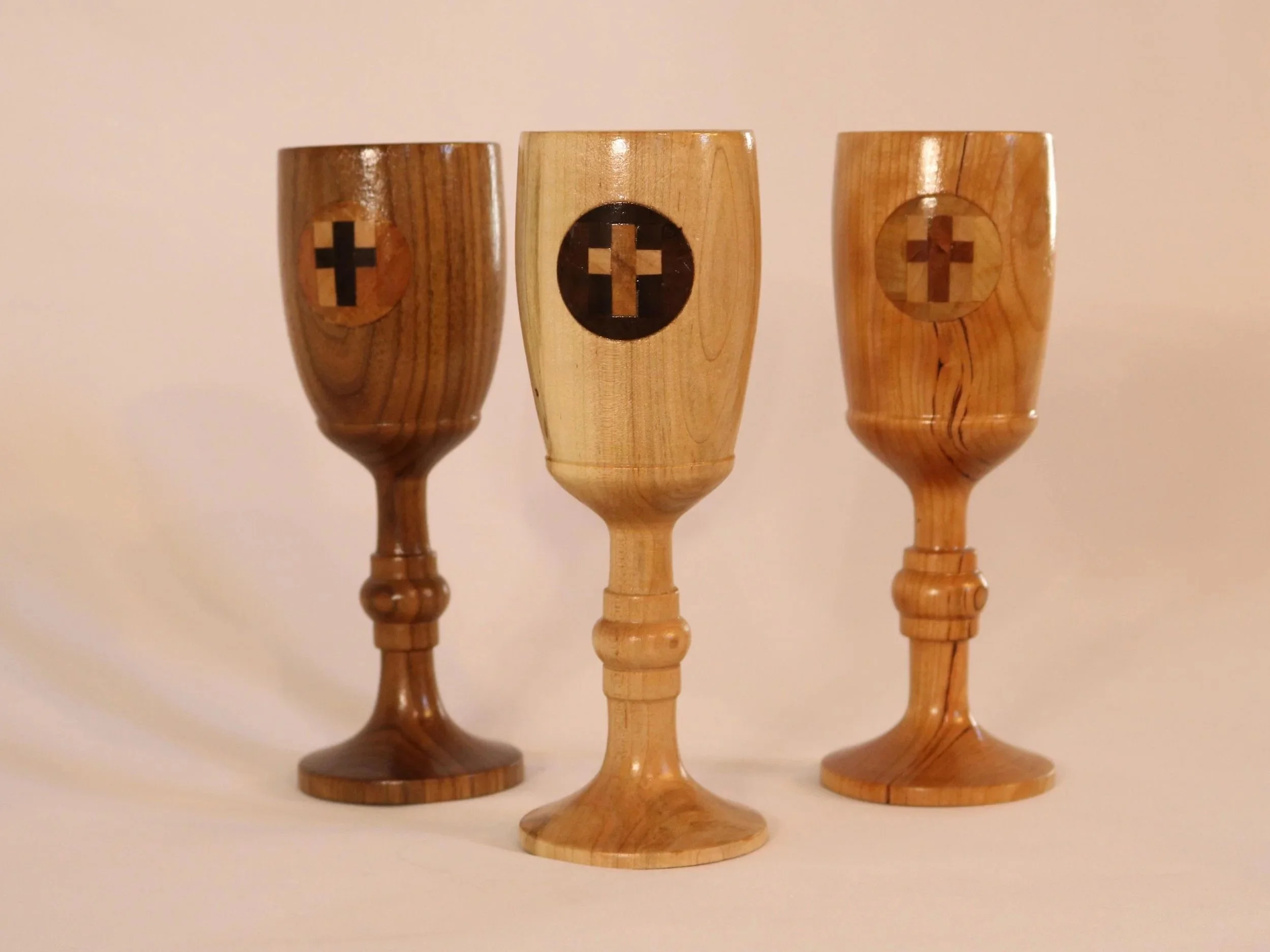 Three wooden chalices with crosses on circular plaques, arranged in a row against a plain background.