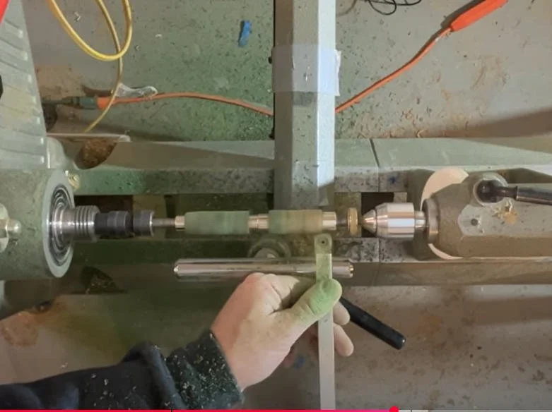A person using a lathe machine to shape a metal rod, wearing a glove, with various tools and cords in a workshop environment.