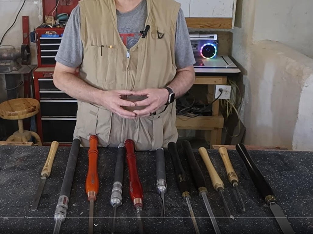A person standing behind a workbench displaying various chisels with wooden and metal handles, in a workshop setting.