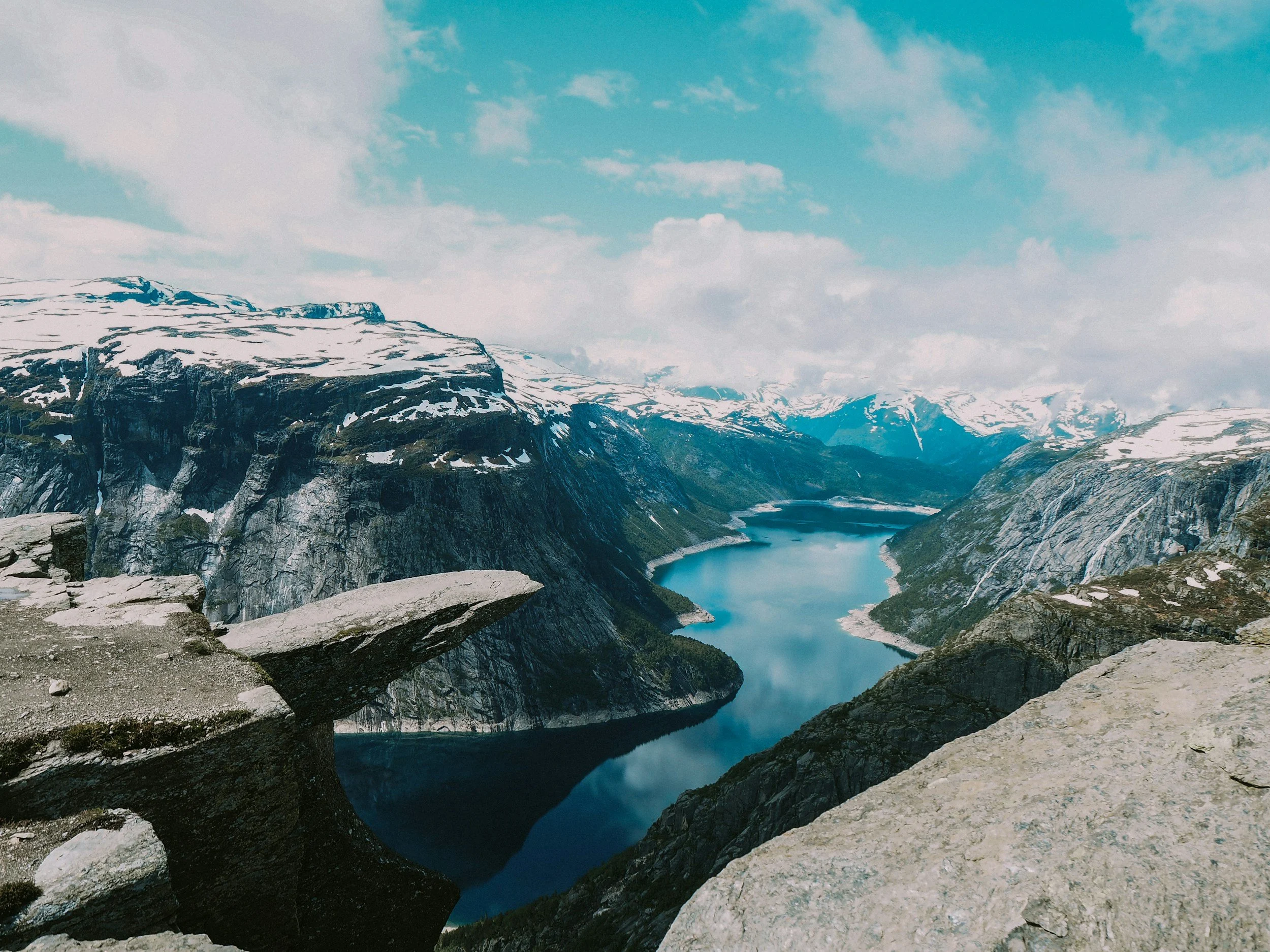 A scenic view of a fjord surrounded by snow-capped mountains and rocky cliffs under a partly cloudy sky.