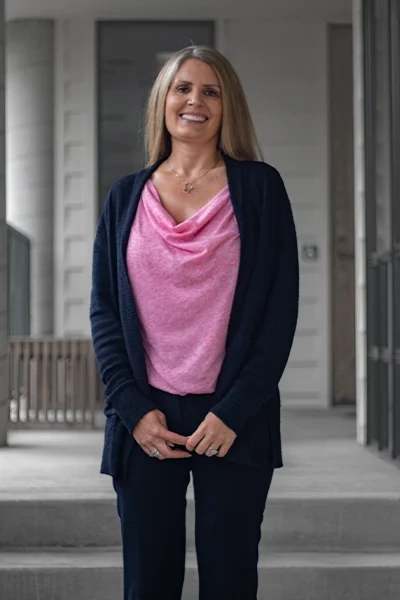 A smiling woman with shoulder-length blonde hair standing outdoors in front of a house, wearing a pink top and navy blue cardigan and pants.