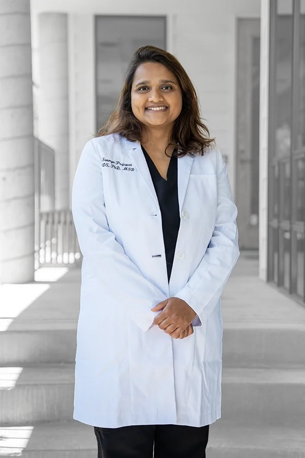 A smiling woman in medical scrubs and a white lab coat standing outside a modern building.