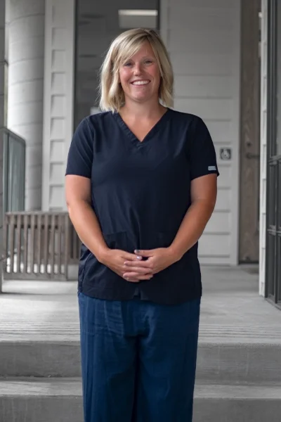 A woman with blonde hair in blue scrubs standing outside a house with a porch, smiling at the camera.