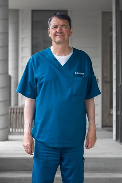A middle-aged man in blue medical scrubs standing outside a building.
