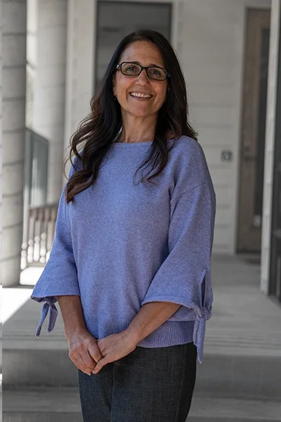 A woman with long dark hair and glasses smiling, wearing a light purple sweater with tied sleeves, standing outdoors in front of a building.