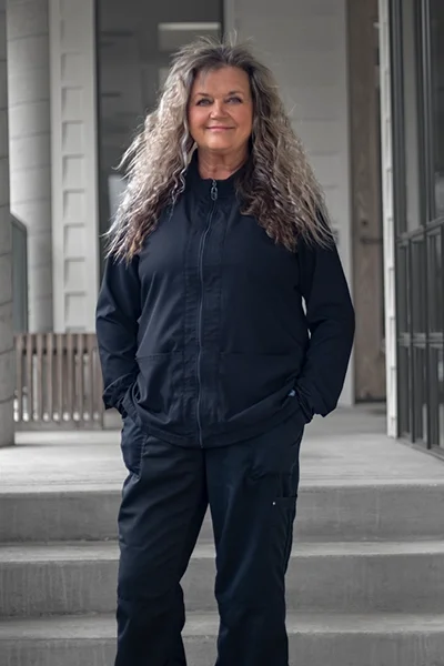Middle-aged woman with long, gray, curly hair standing outside a modern building in a black sports jacket and matching pants, smiling.