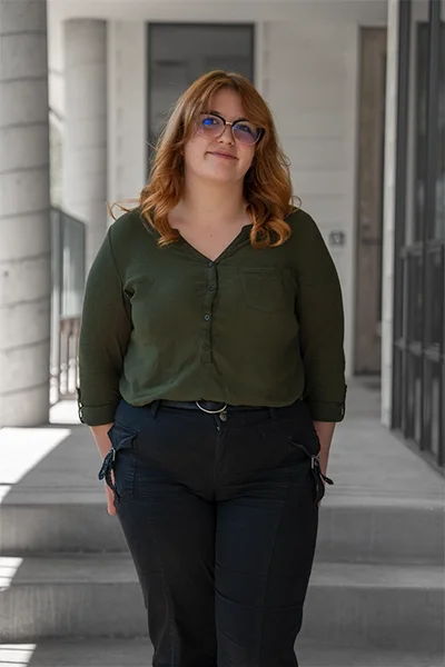 Woman with red hair, glasses, wearing a green shirt and black pants, standing on a modern outdoor walkway.