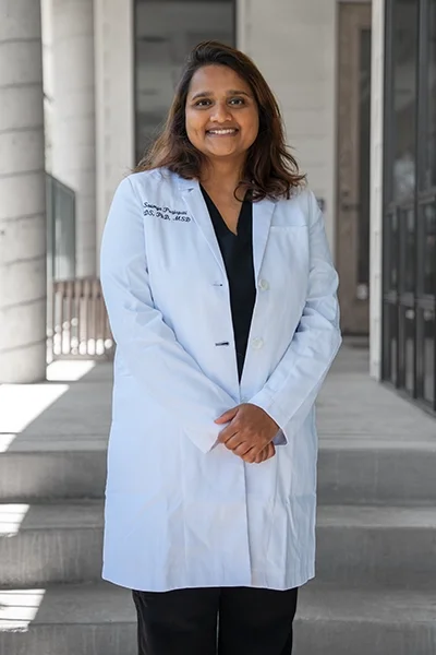A woman in a white lab coat standing outside near a modern building, smiling at the camera.