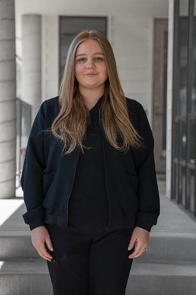 Young woman with long light brown hair wearing a black jacket and black pants standing outside modern building.