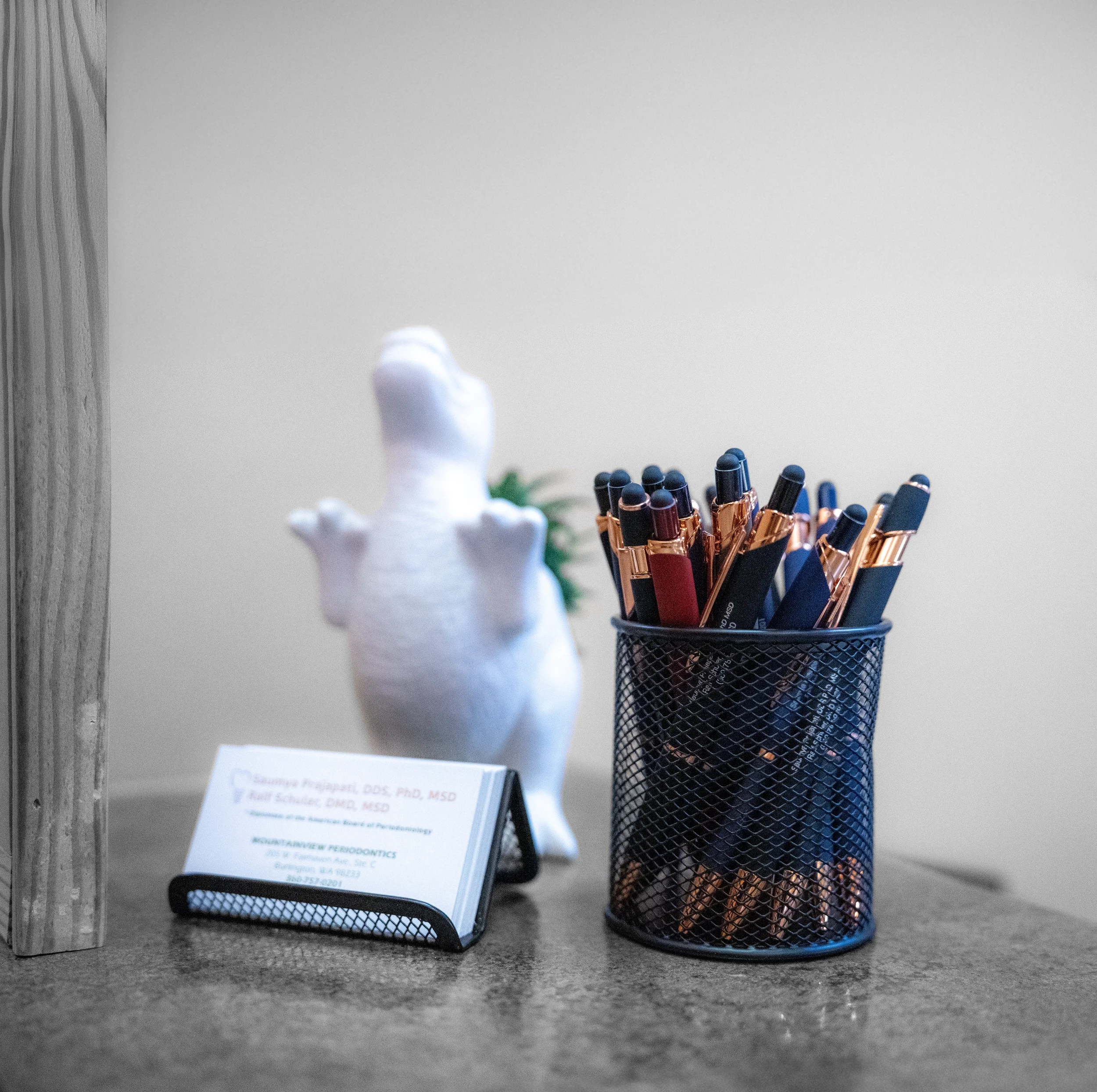 Office desk with a small business cardholder, a black mesh pen holder filled with black and blue pens with copper accents, a white plush toy, and a small green plant. The desk surface is gray and textured, with a wooden vertical surface on the left background.
