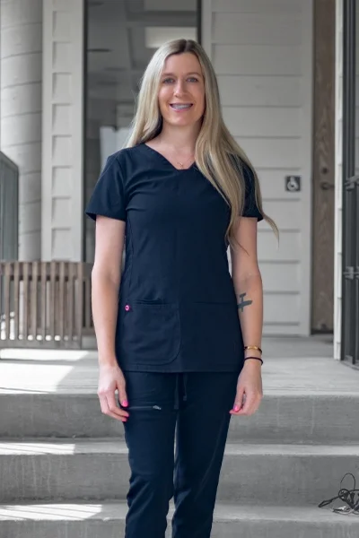 A smiling woman in navy scrubs standing outside a building with steps and a wooden porch.
