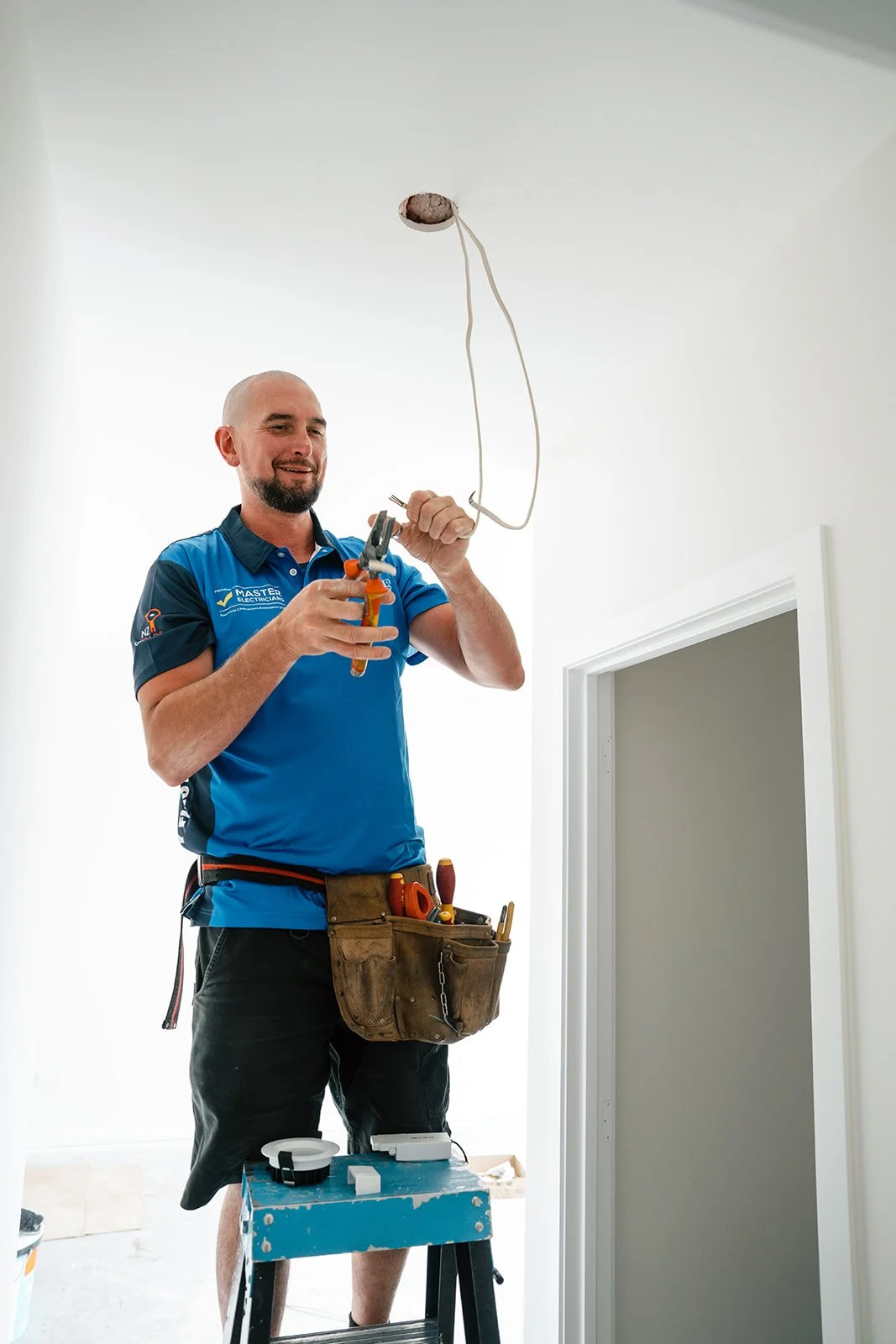 A electrician working on wiring in a ceiling, standing on a step stool, wearing a tool belt with various tools.