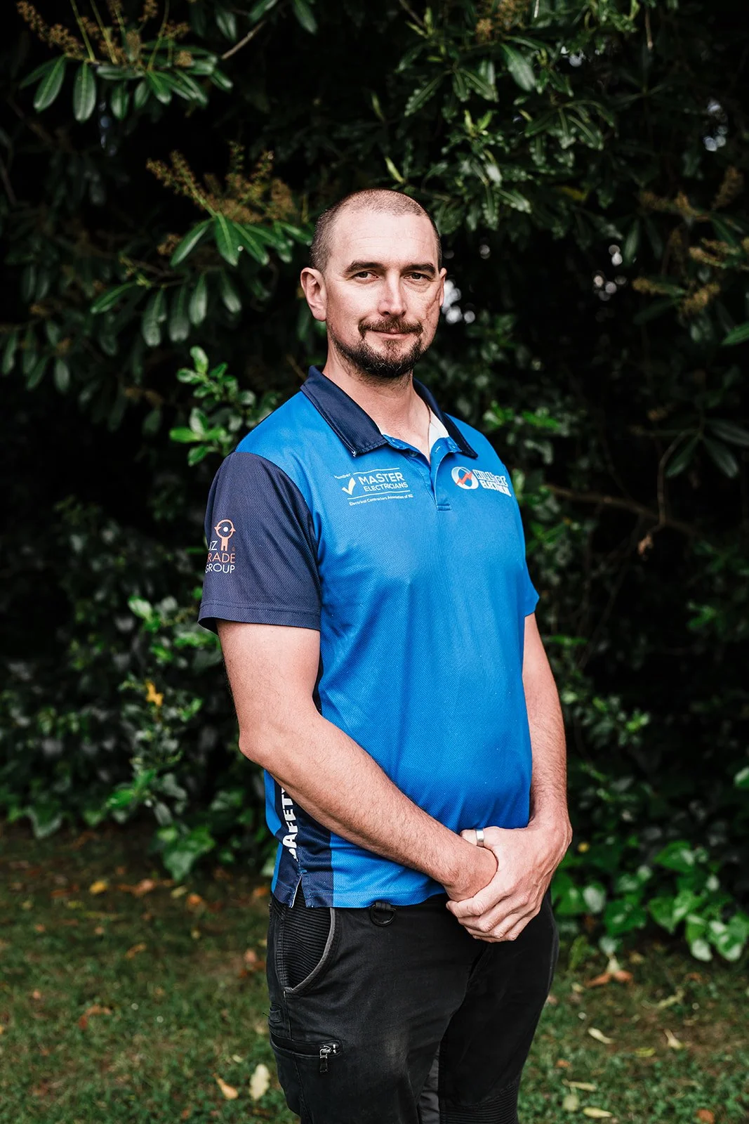 A man wearing a blue sports polo shirt standing outdoors in front of greenery, with short hair, a beard, and smiling slightly.