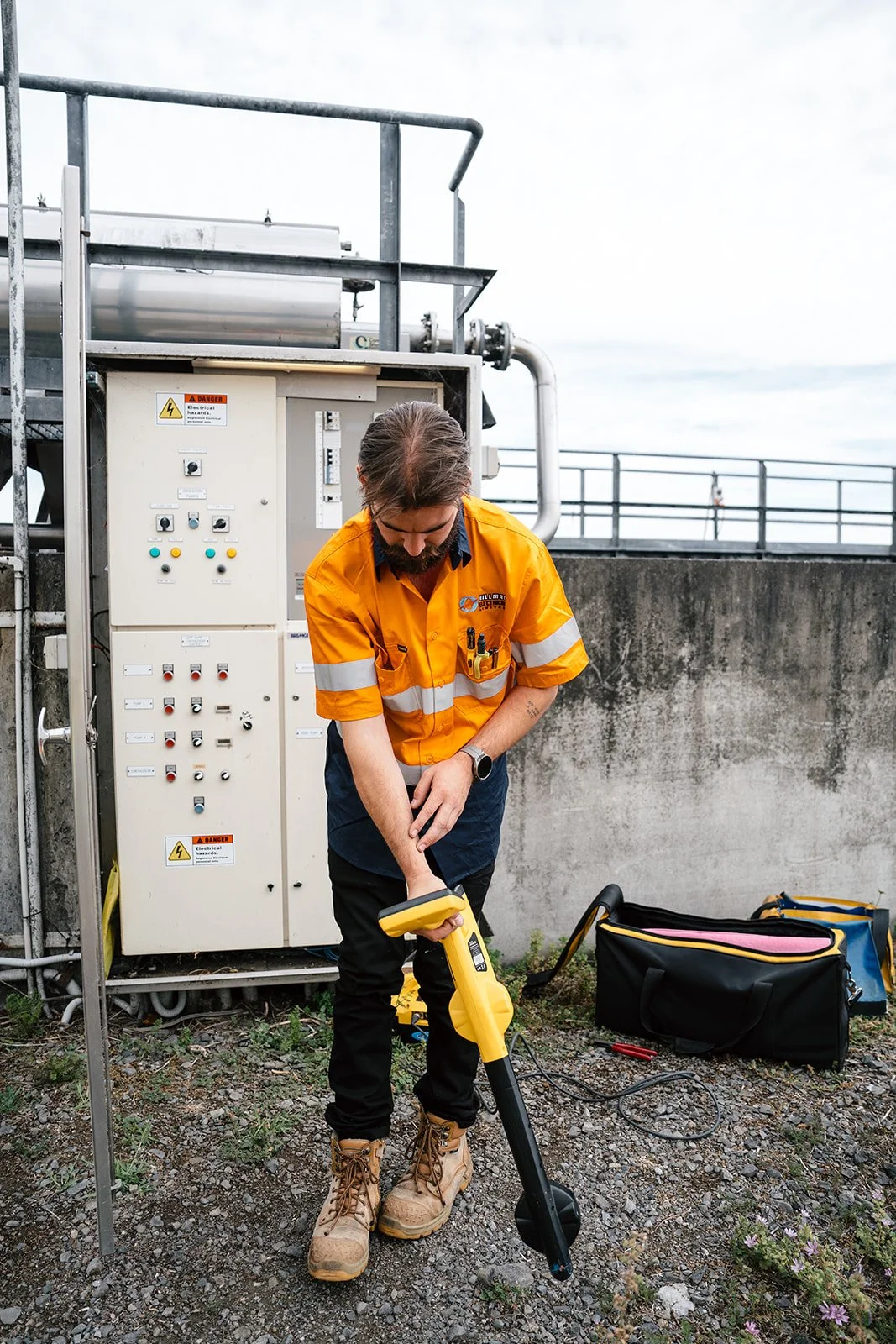 A worker in an orange safety shirt and tan boots using a yellow handheld device outdoors near electrical equipment.