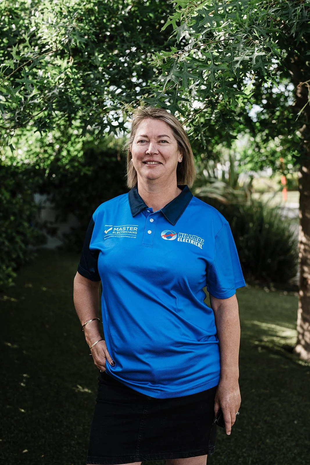 A woman in a blue polo shirt standing outdoors under a tree, smiling at the camera.