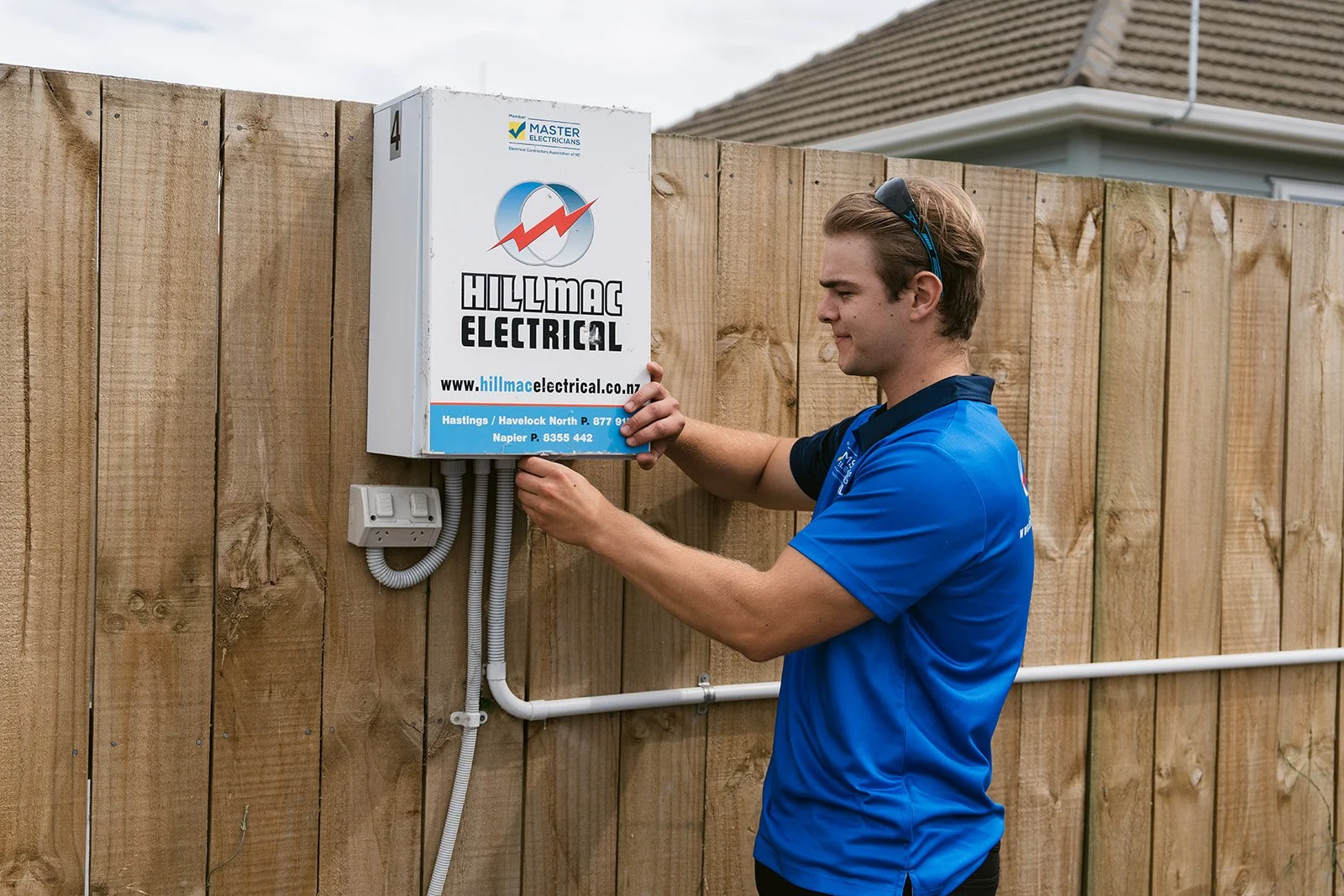 A man in a blue shirt installing an electrical box on a wooden fence.