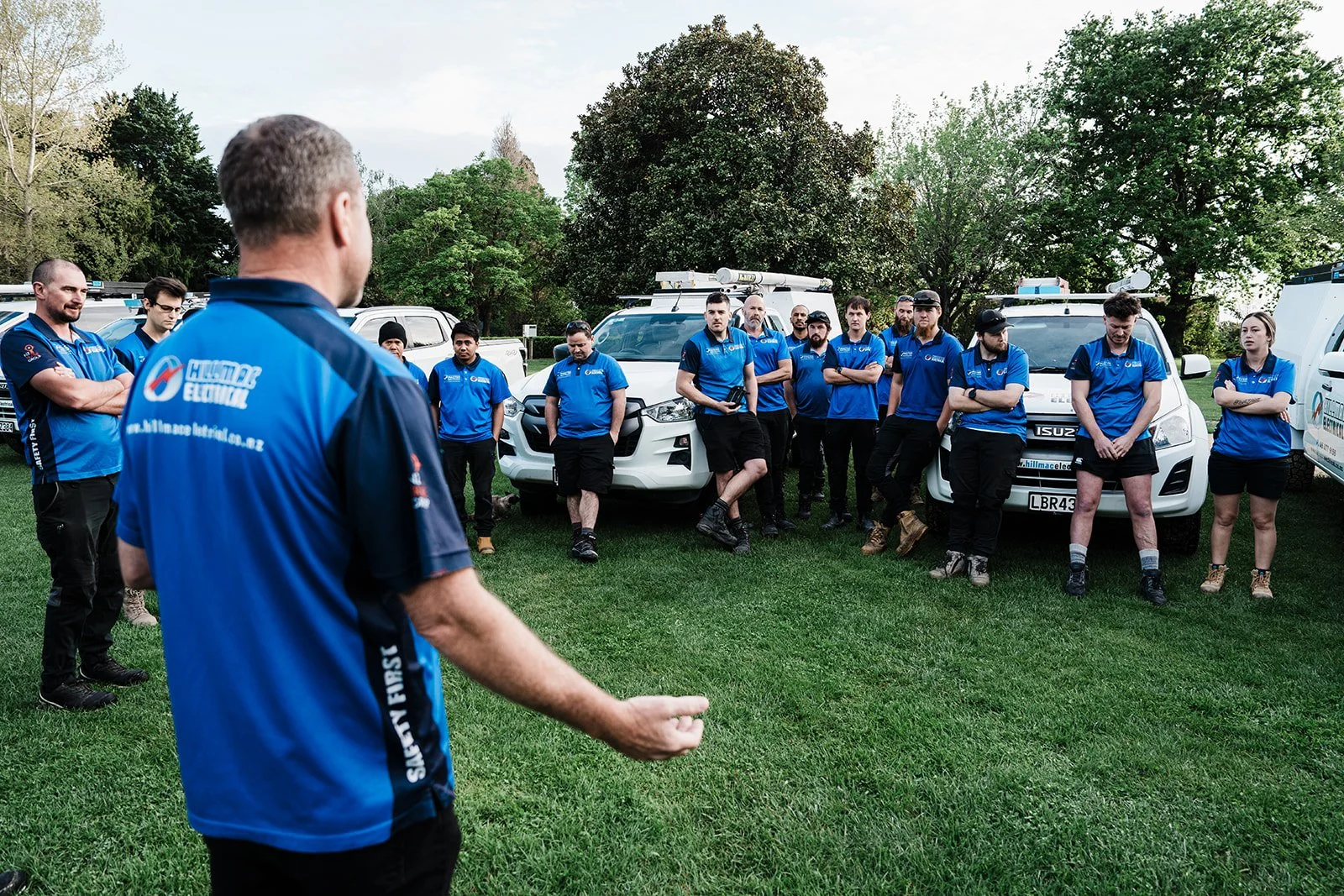 A group of people in blue uniforms gathering outdoors on a grassy area, listening to a person who is speaking, with green trees and parked white vehicles in the background.
