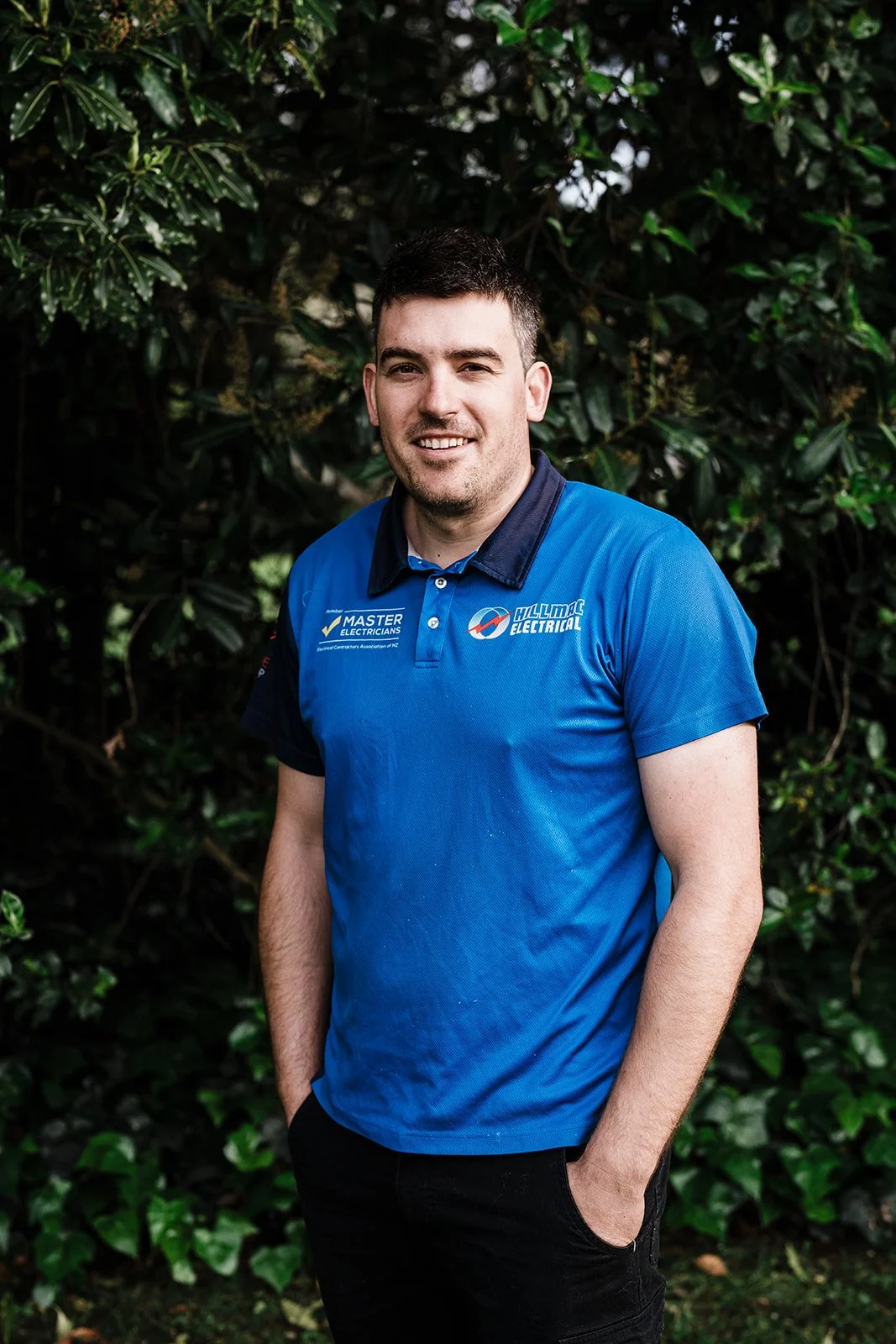 A young man with short dark hair, light skin, and a slight beard, smiling, standing outdoors in front of green leafy bushes. He is wearing a blue polo shirt with logos on it, and his hands are in his pockets.