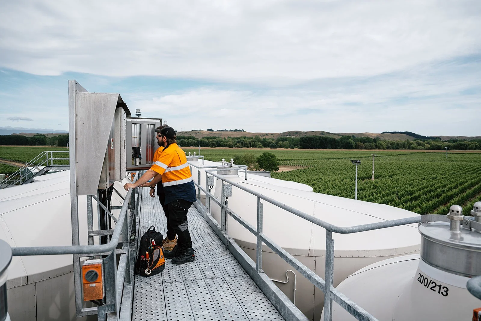 A man in an orange and navy safety vest and black hard hat inspecting equipment on a rooftop of a large industrial tank system in a rural farmland area with green fields and hills in the background.