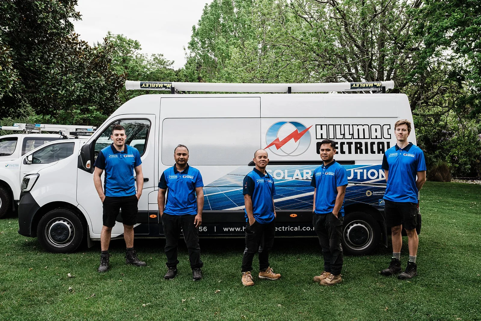 Group of five men standing in front of a white work van with branding for 'HILLMAC ELECTRICAL' and solar panels, outdoors on a grassy area with trees in the background.