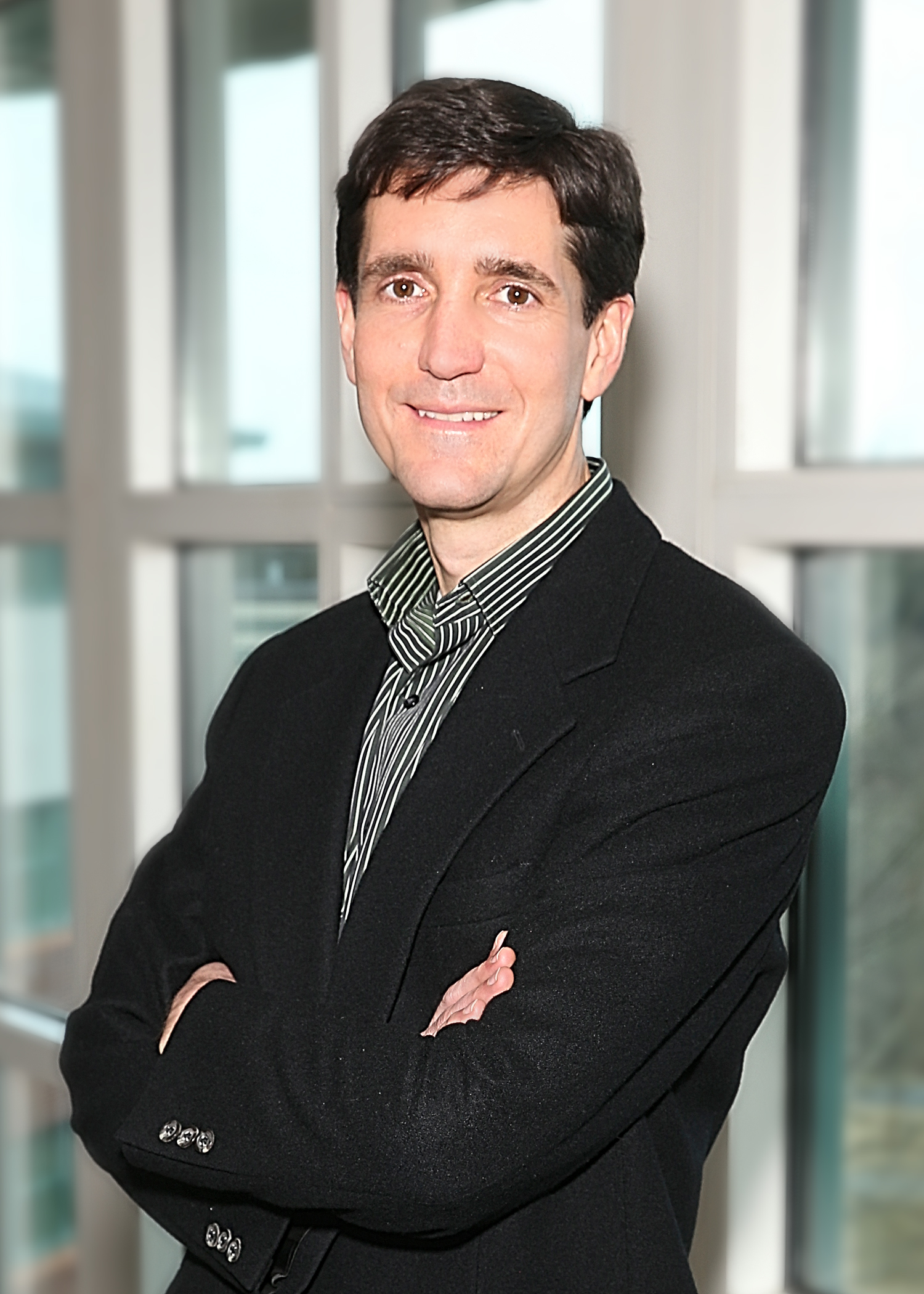 A man with dark brown hair, wearing a black blazer over a striped shirt, standing with arms crossed in front of a large window with blurred cityscape background.