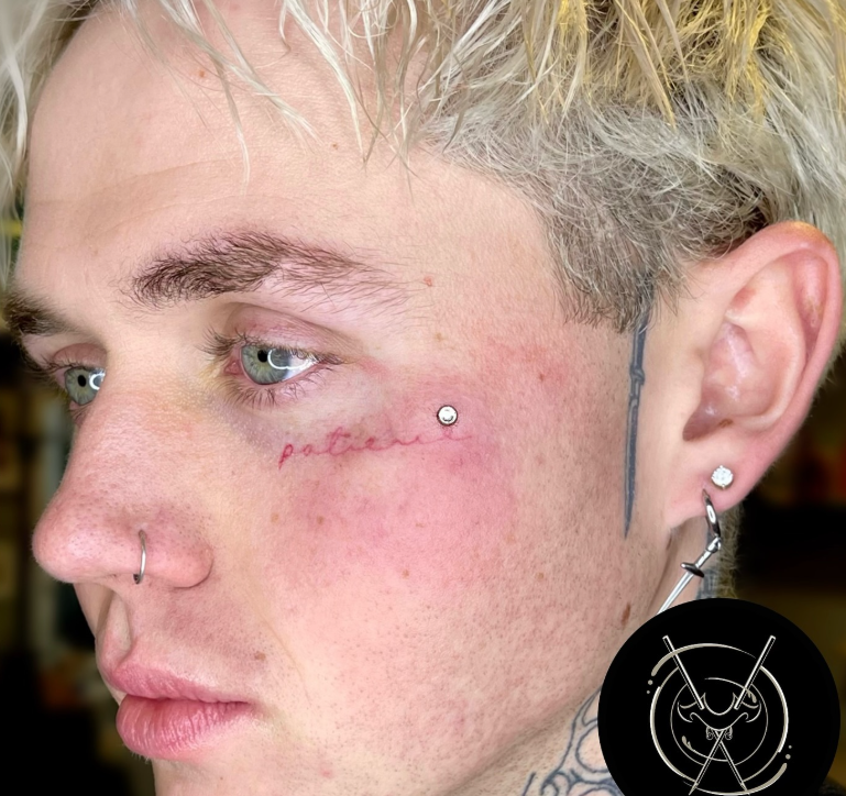 Close-up of a person with blonde hair and multiple facial piercings, including a nose ring, eyebrow piercing, and earrings. They have tattoos on their face and neck, and the word 'patience' tattooed in cursive beneath their eye.