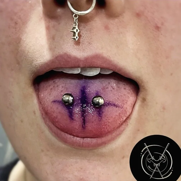 Person with two silver tongue piercings, purple ink tattoo on tongue, wearing a piercing jewelry with a cross charm, and a black circular sticker with lines and symbols.
