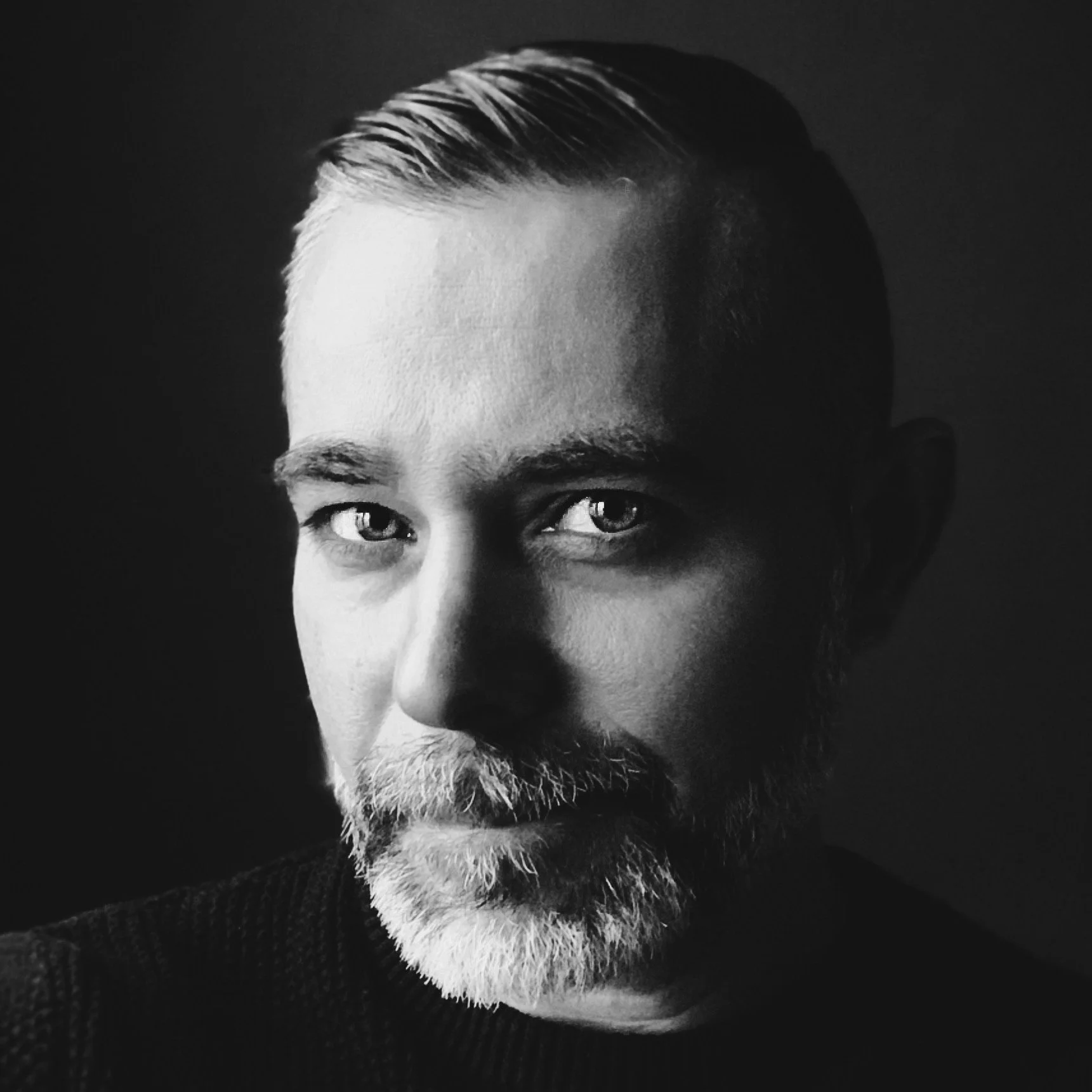 Close-up black and white portrait of a man with light-colored hair and beard, looking directly at the camera.