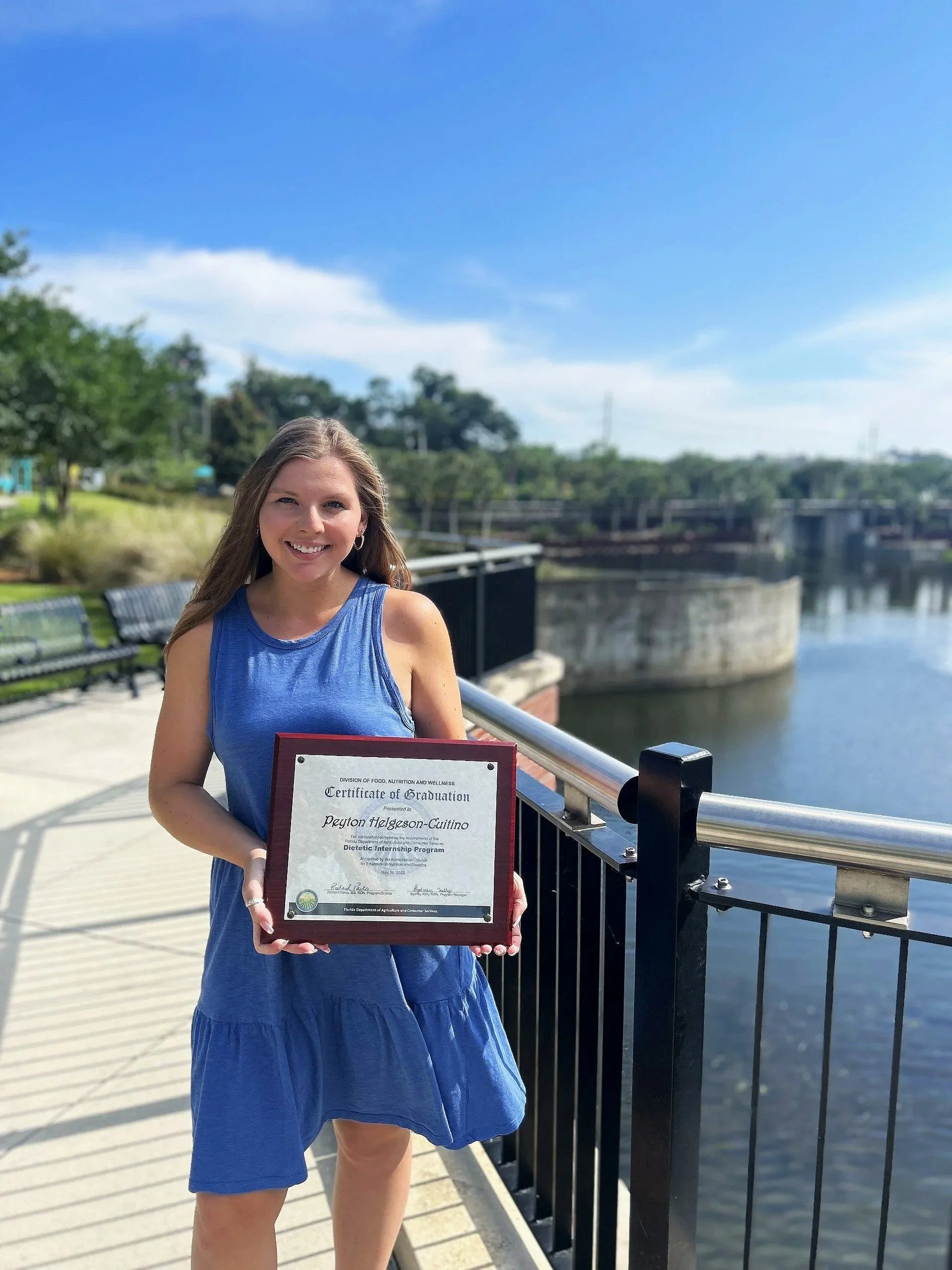 A young woman standing outdoors on a bridge, holding a framed certificate of graduation, smiling at the camera with a river and trees in the background.