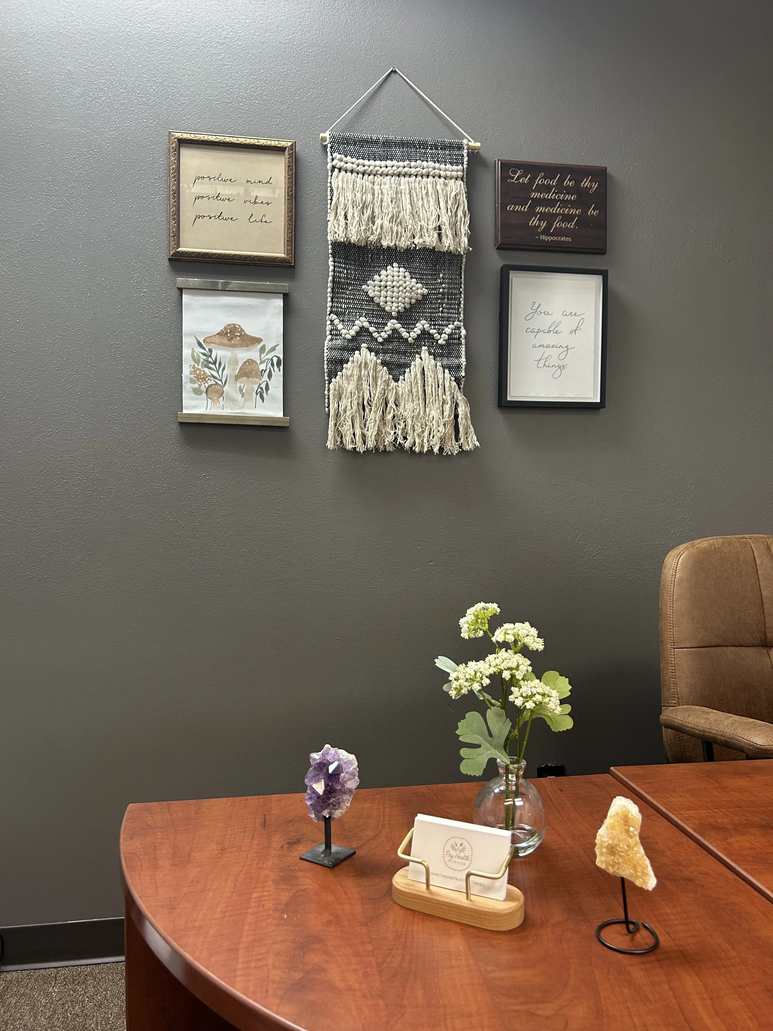 A wooden table with a glass vase holding green and white flowers, a purple crystal cluster, and a decorative mineral specimen. In the background, a gray wall with framed quotes, artwork, and a woven wall hanging.