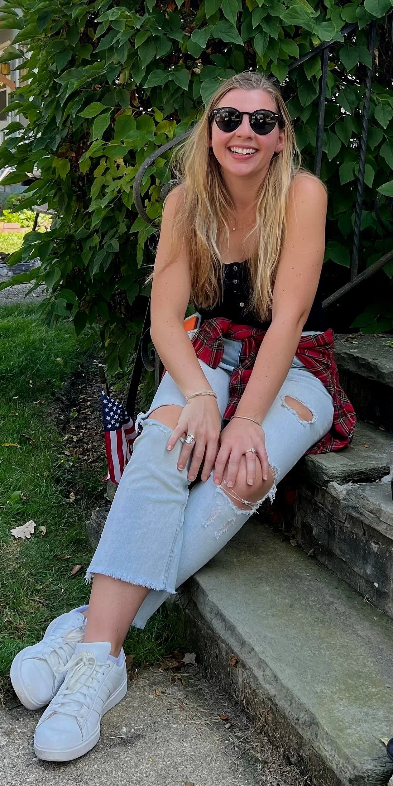 A young woman with long blonde hair wearing sunglasses, a black top, distressed light blue jeans, and white sneakers, sitting on stone steps outdoors in front of green foliage and smiling.