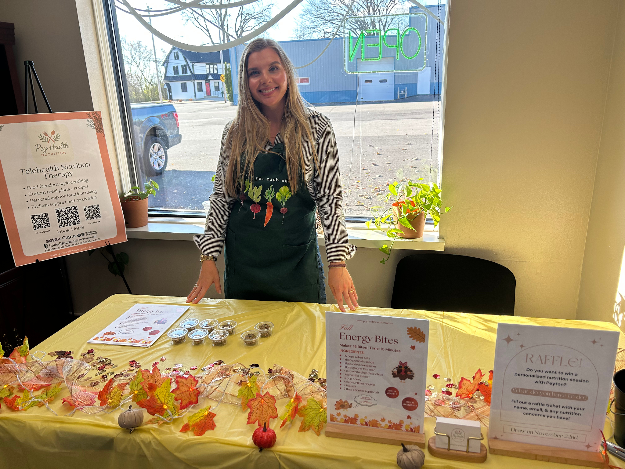 Smiling woman standing behind table with energy bites at a health event, in front of a large window with 'OPEN' sign and outdoor view.