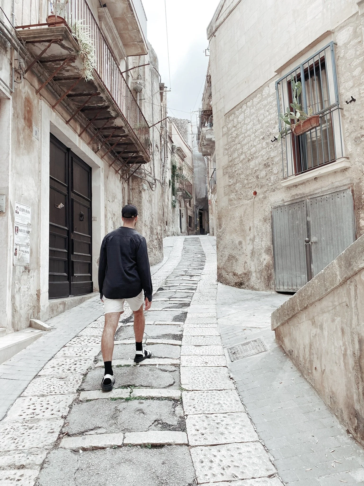 A person walking up a narrow, cobblestone alleyway with old, weathered buildings on each side, featuring balconies and a potted plant in a window.