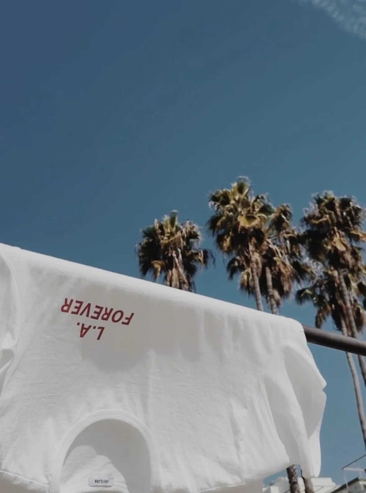 Close-up of a white laundry drying cover with red text that reads "L.A. FOREVER" hanging outdoors against a clear blue sky with tall palm trees in the background.