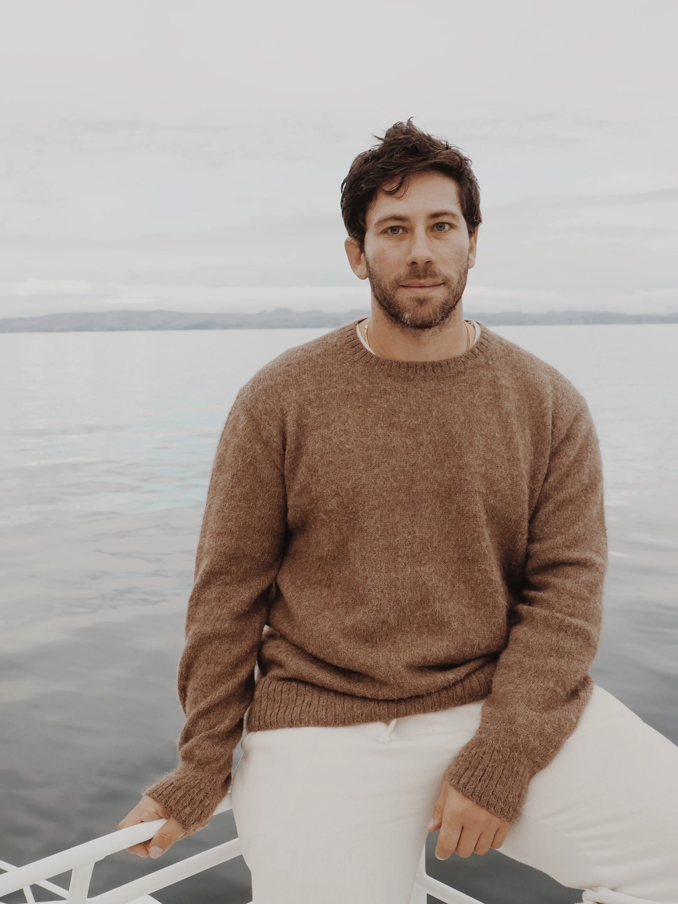 A man sitting on a boat by a body of water with a shoreline in the background, wearing a brown sweater and white pants.