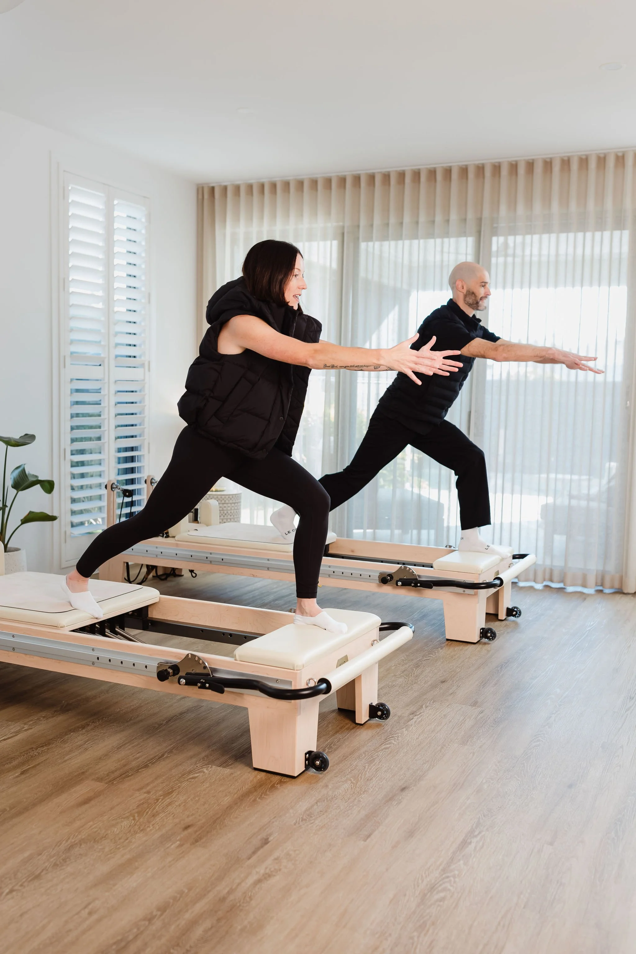 Studio em. Academy Pilates educators Em and Arron demonstrating a standing lunge on Reformer Pilates machines in a bright, contemporary studio.