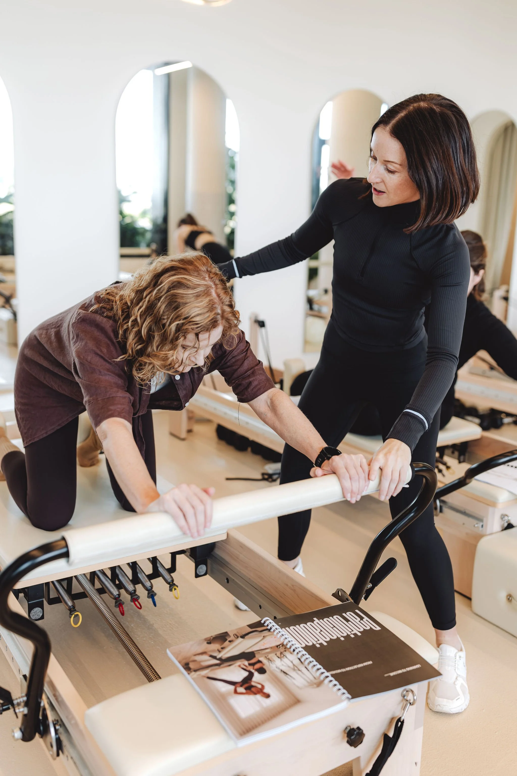 A woman assisting another woman with a physical therapy exercise using a parallel bar in a well-lit fitness or rehabilitation center, with mirrors and other people in the background.