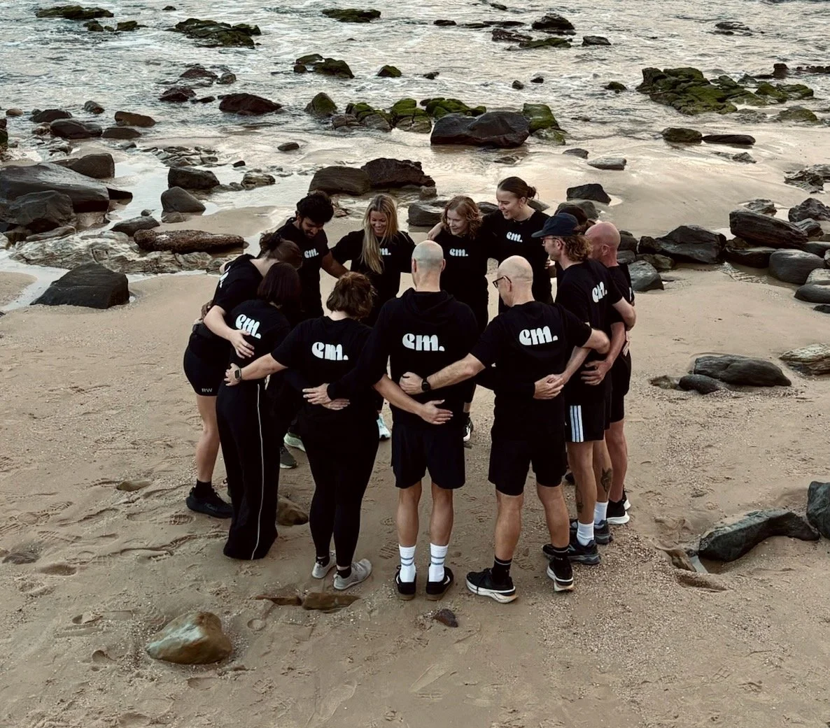 A group of people wearing matching black shirts with 'em.' on the back, standing in a circle on a sandy beach with rocks, near the ocean.