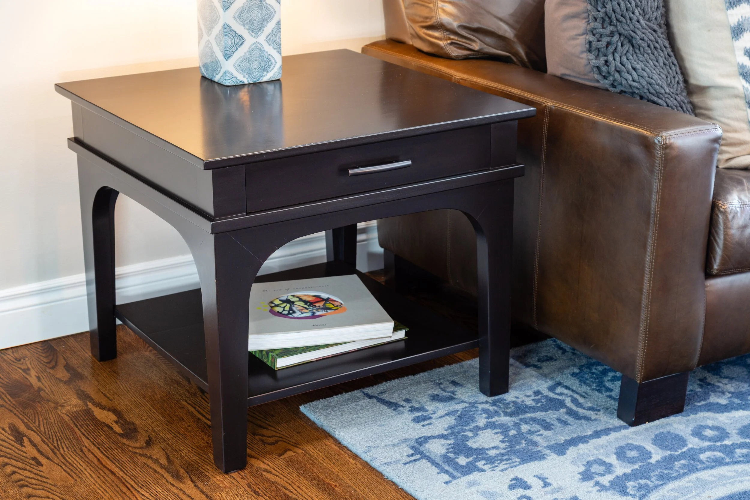 Black wooden side table with a drawer and lower shelf, positioned next to a brown leather sofa with assorted gray and beige cushions, on a blue decorative rug on a wooden floor.