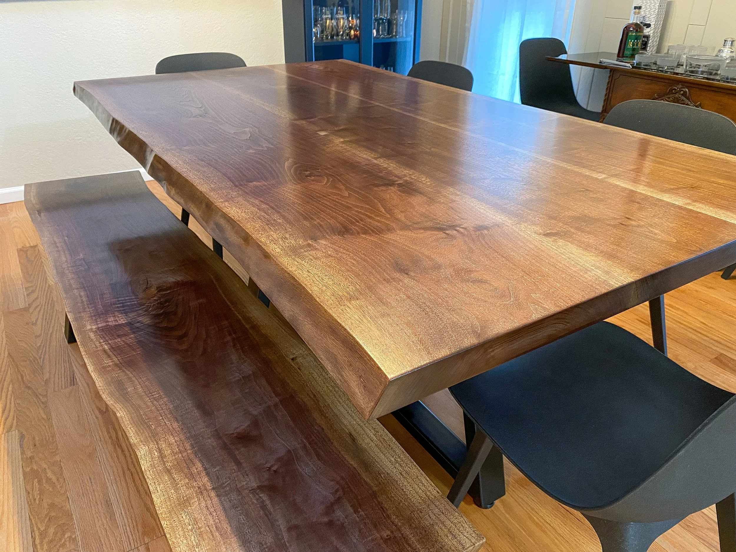 A Walnut Live Edge dining table with a polished surface and a matching bench, situated in a room with wooden flooring and black chairs. A china cabinet filled with glassware is visible in the background.