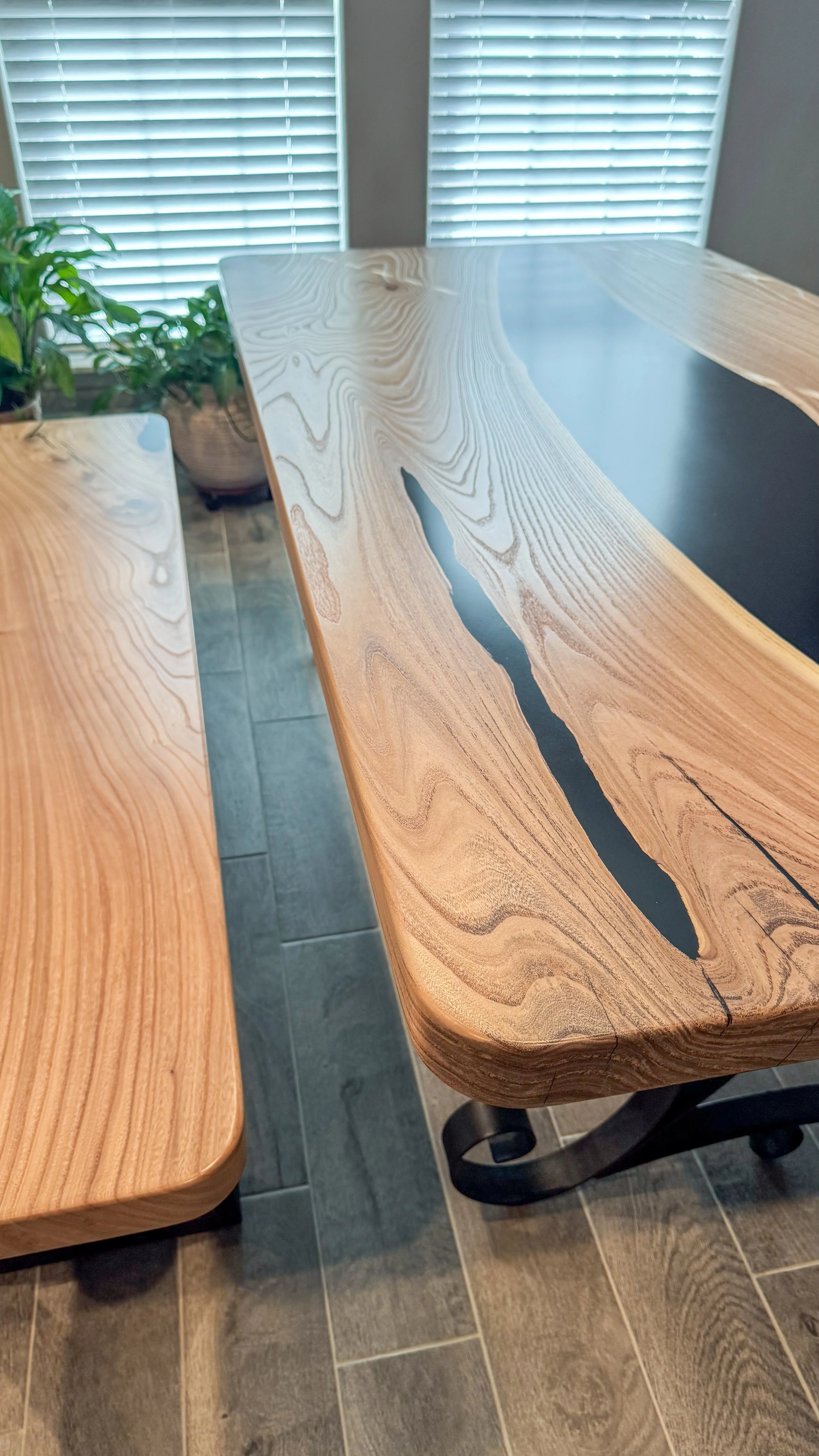 An American Elm Epoxy River table and matching bench positioned next to each other in a room with large window blinds and potted plants in the background.