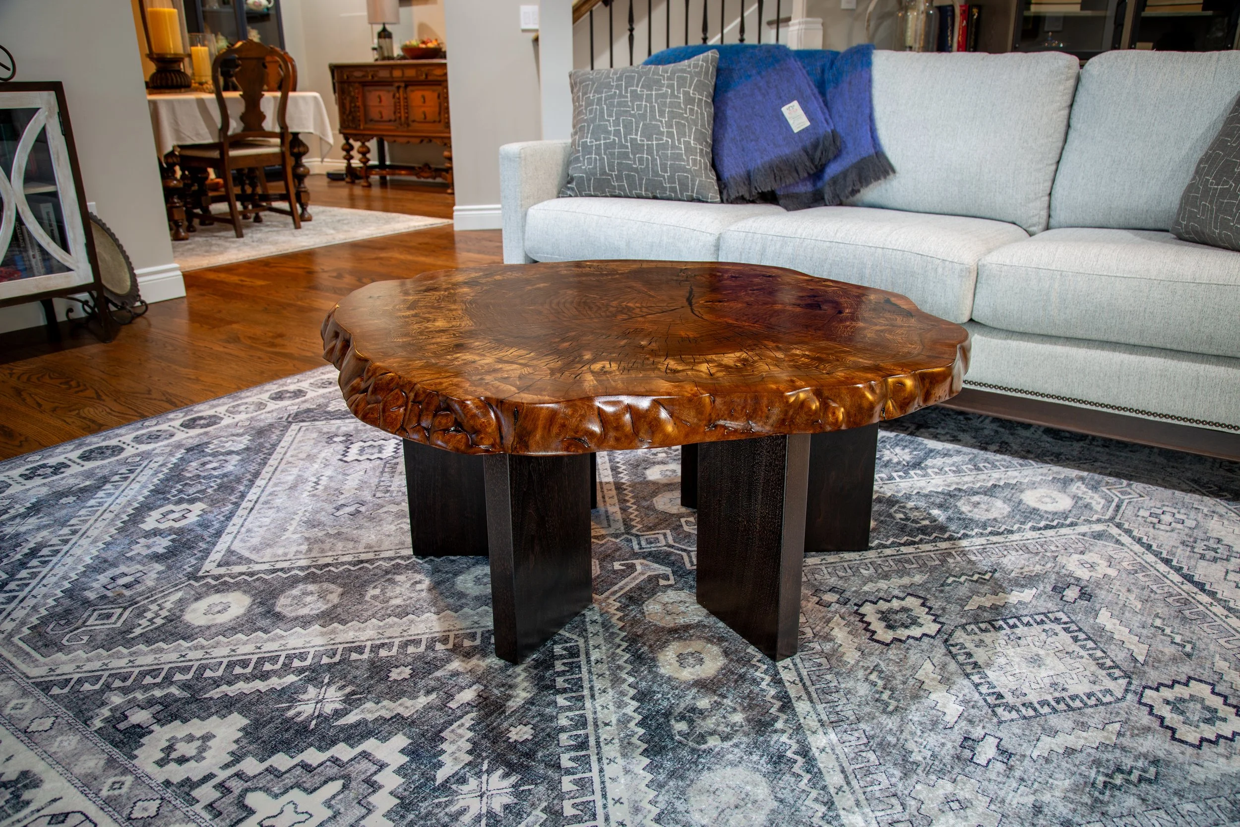 A living room with a Claro Walnut coffee table, a light-colored sofa with decorative pillows and throws, a patterned area rug, and a bookshelf in the background.