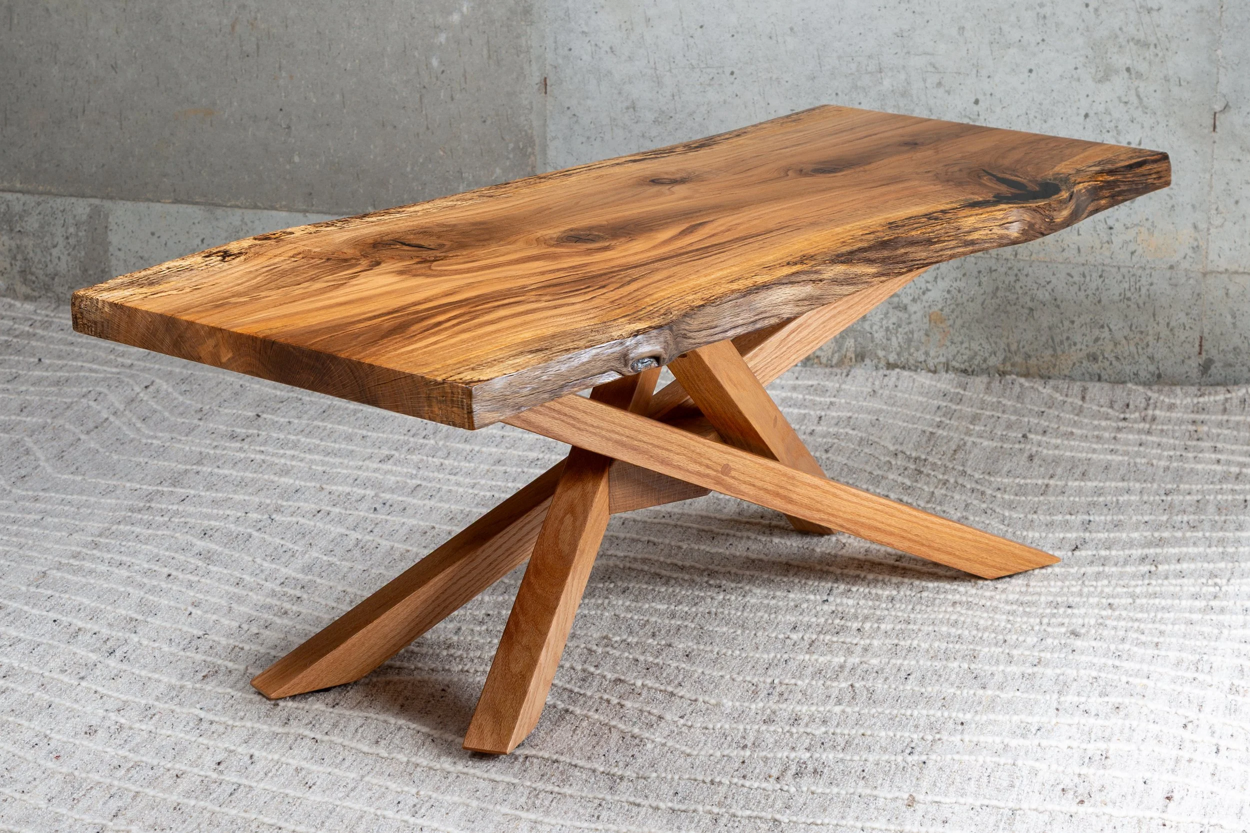 A Live Edge Pin Oak coffee table with intersecting oak legs for the table base placed on a textured gray and cream rug.
